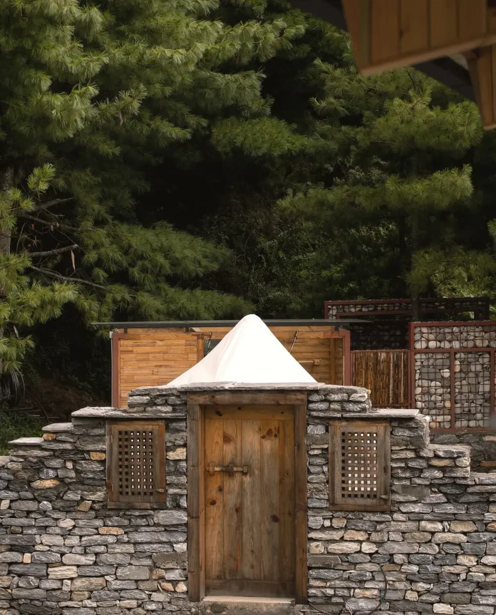 Canvas tent peak rising behind a dry-stone wall and timber gate among the pines