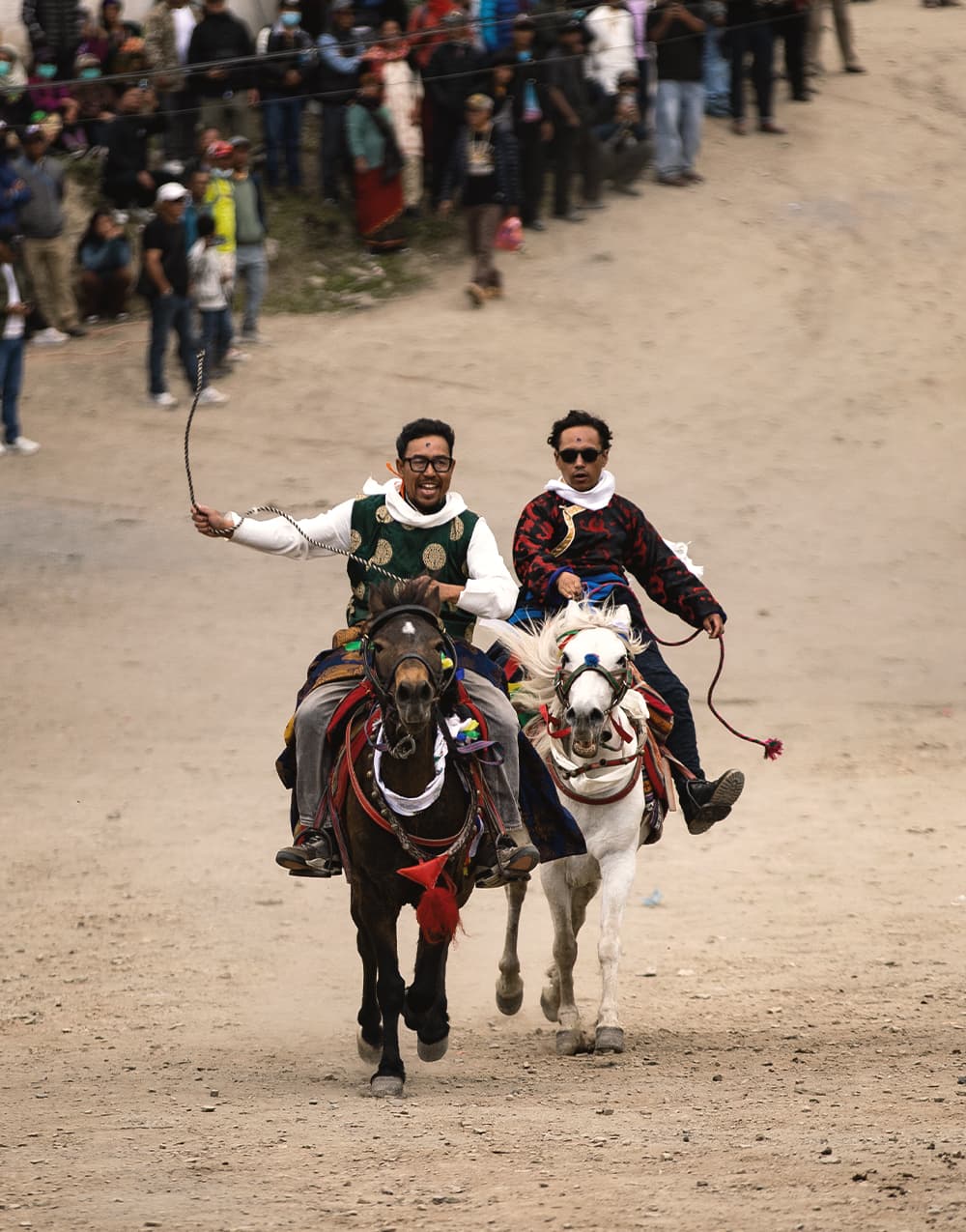 Two riders racing their horses past a crowd at a village festival