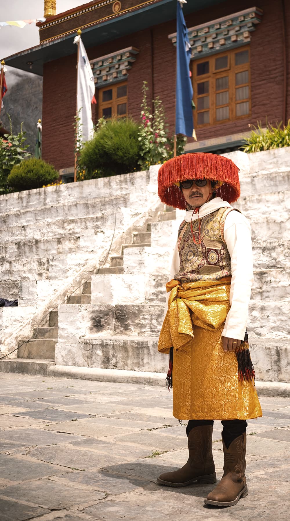Man in a red-fringed festival hat and gold robe outside a monastery