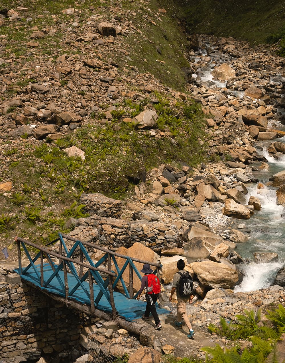 Two hikers crossing a blue timber bridge over a rocky mountain stream