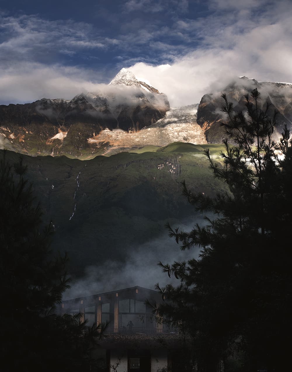 Camp lodge in evening mist below a snow peak and waterfall-streaked ridges